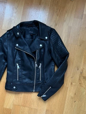 Svart skinnjacka från Vero Moda - Snygg svart skinnjacka med klassisk bikerstil från Vero Moda. Jackan har silvriga dragkedjor, knappar och detaljer, samt breda slag. Passformen är normal och jackan har långa ärmar med dragkedjor vid ärmsluten. Ej riktigt läder. Oanvänd.