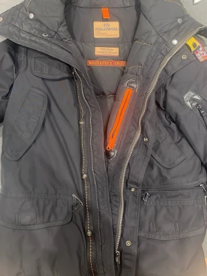 Svart Parajumpers parkajacka - Säljer en svart Parajumpers parkajacka med flera fickor, gul detalj på axeln och orange dragkedja. Jackan har huva, patch på ärmen och robusta dragkedjor. Perfekt för kalla dagar och riktigt snygg streetstil. Pris kan diskuteras