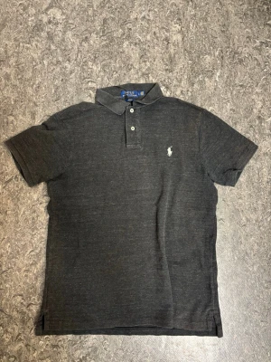 Mörkgrå pikétröja från Polo Ralph Lauren - Säljer en mörkgrå pikétröja från Polo Ralph Lauren i slim fit-modell. Tröjan har klassisk krage, två knappar framtill och den ikoniska broderade loggan på bröstet. Tillverkad i mjuk bomull och har korta ärmar. Perfekt till en clean och stilren look.