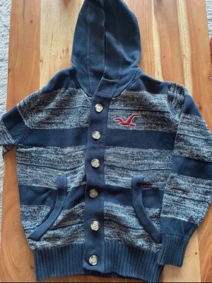Hollistwr cardigan - Mysig marinblå och grå randig hoodiekofta från Hollister med stora knappar framtill och broderad logga på bröstet. Tröjan har huva, två fickor och är stickad i mjukt material. Perfekt för kyliga dagar och enkel att matcha.