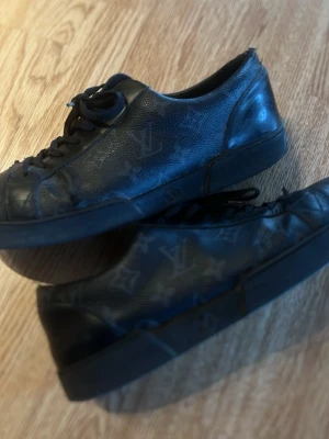 Svarta Louis Vuitton sneakers i skinn - Säljer ett par svarta Louis Vuitton sneakers i skinn med klassiskt LV-monogram över hela skon. Skorna har rund tå, svarta snören och platt sula. Perfekta för dig som gillar exklusiva detaljer och streetstyle. Tydligt slitage finns fram innan toebox och även i hälen på insidan av skon, därav priset. Kvitto från KASPERSHEAT finns även. 