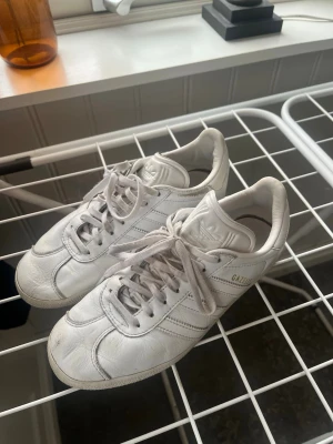Vita Adidas Gazelle sneakers - Säljer ett par klassiska vita Adidas Gazelle sneakers i skinn. Skorna har snörning och låg profil, perfekt till jeans eller shorts. Väl använda så pris kan diskuteras!