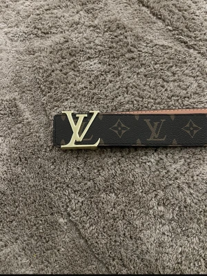 Brun monogram bälte - Säljer ett klassiskt brunt  bälte med ikoniskt monogrammönster och stor logga i guld som spänne. Perfekt accessoar för att lyfta vilken outfit som helst.