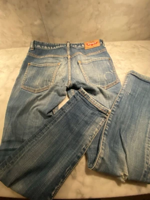 Blå jeans från Dsquared2 - Säljer ett par klassiska blå jeans från Dsquared2 med slitningar och snygg tvätt. Jeansen har fem fickor, gylf med knappar och detaljerade sömmar. Perfekt för dig som vill ha en avslappnad men ändå trendig look. Passformen är original Slim-fit 