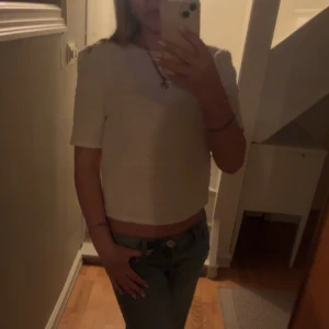 Vit kortärmad topp med guldknappar H&M - Säljer en vit, strukturerad topp från H&M med korta ärmar och croppad passform. Toppen har tre dekorativa guldknappar på ena axeln och är gjord i ett tjockare, vävt material som ger en exklusiv känsla. Perfekt till jeans eller kjol för en trendig look.