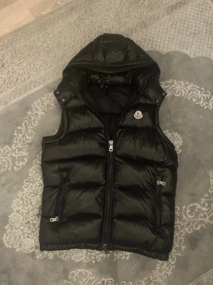  Moncler väst  - Säljer en svart dunväst från Moncler med avtagbar huva och två dragkedjeförsedda fickor. Västen har quiltad design, hög krage och Moncler-logga på bröstet. Perfekt för lager-på-lager och riktigt snygg till höst- och vinteroutfits.