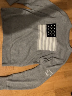 Grå stickad tröja från GANT - Säljer en grå stickad tröja från GANT med amerikansk flagga i vitt och svart på bröstet och broderad text 'GANT NH-CT 1949' på ena ärmen. Tröjan har rund hals, ribbade muddar och lång ärm. Perfekt för dig som gillar klassisk college-stil.