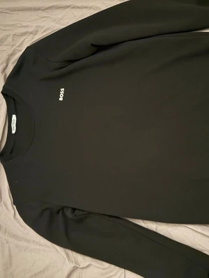 Boss tröja - Svart långärmad tröja från BOSS med enkel och clean design. Tröjan har rund halsringning och en liten vit BOSS-logga på bröstet. Perfekt för dig som gillar stilrena plagg med premiumkänsla.