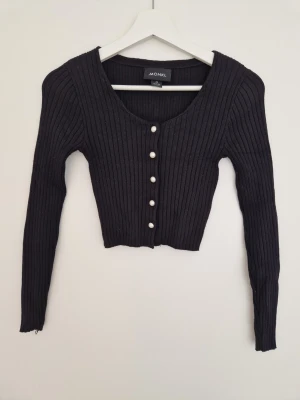Svart ribbad kofta från Monki XS - Säljer en svart ribbad kofta från Monki i storlek XS. Koftan är croppad med långa ärmar och har snygga pärlknappar framtill. Tillverkad i mjuk viskosmix som känns skön mot huden. Perfekt till lager på lager och enkel att styla. Blivit lite smått nopprig i tyget, men fortfarande fin.