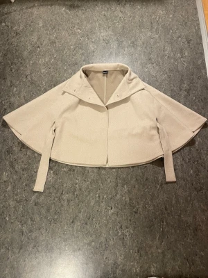 Beige capejacka från Gina Tricot - Säljer en trendig ponchojacka från GinaTricot. Knappt använd och i superbra skick. Storlek XS/S och jätteskön, perfekt för sen vinter/ vår. Säljer för 400kr.