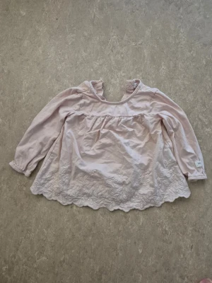 Blus - Säljer en söt ljusrosa blus från Newbie i storlek 74. Blusen har långärmade ärmar med resår, hals med knapp och fina broderade spetsdetaljer längs nederkanten. Perfekt för dig som gillar romantisk och mjuk stil.
