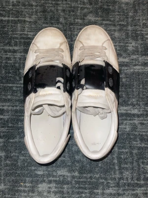Valentino Garavani Open sneakers vit/svart - Säljer ett par Valentino Garavani Open sneakers i vitt skinn med bred svart läderdetalj över snörningen. Klassisk rund tå och platt sula, med vita skosnören och subtilt guldtryck på insidan. Snygg kontrast mellan färgerna och exklusiv känsla. Pris kan diskuteras vid snabb affär.