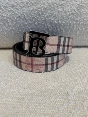 Burberry bälte - Snyggt Burberry bälte i skinn med klassiskt rutigt mönster i beige, svart och rött på ena sidan och mörkt rutigt mönster på andra sidan. Bältet har en stor metallspänne med Burberrys ikoniska TB-logga. Perfekt accessoar för att lyfta din outfit. Man kan vända på det så att det blir 2 färger, svart och beige , kom dm för eventuella frågor 🧐 pris kan också diskuteras 💯.