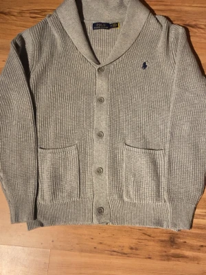 Grå ribbad kofta från Polo Ralph Lauren - Säljer en grå ribbad kofta från Polo Ralph Lauren med klassisk sjalkrage, knappar framtill och två stora fickor. Diskret logga broderad på bröstet. Perfekt för lager på lager under höst och vinter.