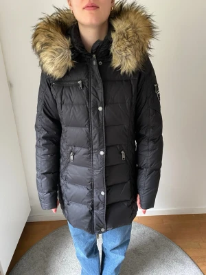 Svart Rockandblue dunjacka med fuskpäls - Säljer en svart dunjacka från Rockandblue i toppskick. Huvan är avtagbar och fuskpälsen kan också tas av separat om man vill behålla huvan på. Jackan har quiltade partier, flera dragkedjeförsedda fickor, två innerfickor och Rockandblue-logga på ärmen. Perfekt för kalla dagar och riktigt snygg till både jeans och finkläder. Storleken är 40 men jag tycker den är mer som storlek 38.