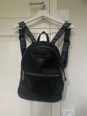 Svart ryggsäck från Guess - Säljer en svart ryggsäck från Guess med justerbara axelband där loggan står tryckt. Väskan har guldfärgade dragkedjor och är gjord i syntetmaterial med struktur. Invändigt är fodret ljust med Guess-mönster. Perfekt storlek för vardagens essentials. Sparsamt använd men sliten vid sömmen, se bild.