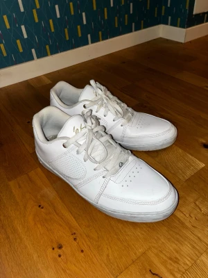 Vita sneakers från éS med snörning - Säljer ett par klassiska vita sneakers från éS med lågt skaft och snörning. Skorna har perforerade detaljer på sidorna och tåpartiet samt guldfärgade detaljer vid snörhålen. Yttermaterialet är i syntet/skinn och sulan är vit med ett grönt éS-märke undertill.