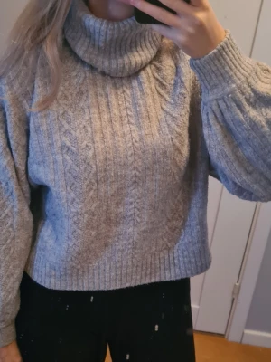 Grå polotröja från H&M Divided - Mysig grå polotröja från H&M Divided i storlek S. Tröjan är stickad med kabelmönster, har en hög polokrage och långa ärmar med ribbade muddar. Perfekt för kyliga dagar och enkel att styla till olika outfits.