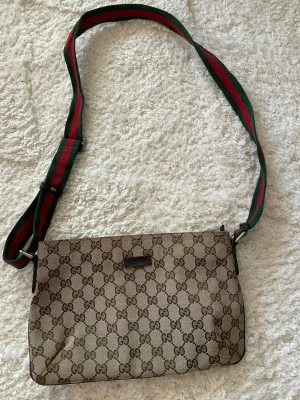 Gucci axelväska med GG-mönster - Säljer en klassisk Gucci axelväska i beige och brunt med ikoniskt GG-mönster. Väskan har en justerbar axelrem i grönt och rött, dragkedja upptill och detaljer i skinn. Perfekt för dig som vill ha en snygg och tidlös accessoar från Gucci.