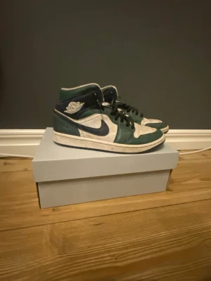 Nike Air Jordan 1 Mid grön/vit - Tja, Säljer ett par Nike Air Jordan 1 Mid i grön och vit färg med svarta detaljer och svarta snören. Skorna har klassisk Jordan-logga på sidan,  man får med org snören också,hör av er!