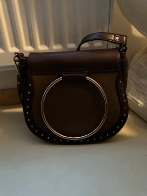 Mörklila axelväska med metallring - Snygg mörklila axelväska i skinn med stor silverfärgad metallring framtill och små nitar längs kanterna. Väskan har justerbar axelrem och praktisk ficka på baksidan.