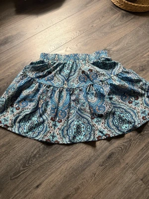 Blå paisleymönstrad kortkjol - Supersnygg kortkjol med paisleyinspirerat mönster i blått, brunt och vitt. Kjolen har resår i midjan och volangdetalj som ger en avslappnad och trendig look. 