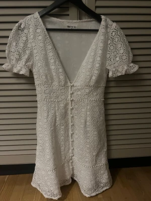 Vit spetsklänning från The Sky - Superfin vit kortklänning i spets med blommigt mönster och v-ringning. Puffiga kortärmar med volangkant och knappar framtill. Perfekt för sommarens alla tillfällen. Sitter snyggt och har en feminin passform.