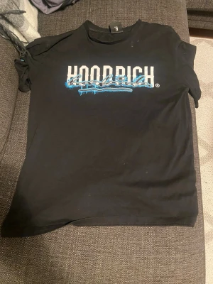 Svart Hoodrich t-shirt - Svart t-shirt från Hoodrich i storlek M med stort vitt och blått tryck fram och bak. Texten 'Hoodrich' och 'From Nothing To Something' ger streetwear-vibe. Klassisk passform och rund hals. Perfekt för dig som gillar snygga statement-plagg.