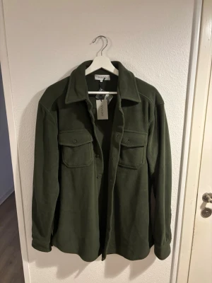 Helt ny grön overshirt från Sandö, stl 50 - Säljer en stilren grön overshirt från Sandö i storlek 50. Jackan har två bröstfickor med knappar, klassisk krage och stängs med knappar framtill. Perfekt lager-på-lager-plagg i mjukt material som funkar till många looks.