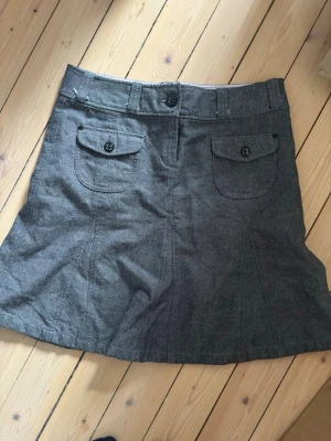 Grå kortkjol från Esprit - Säljer en grå kortkjol från Esprit med två fickor framtill som har lock och knappdetaljer. Kjolen har bälteshällor och stängs med knapp och dragkedja. Materialet känns mjukt och är troligtvis bomull eller en bomullsblandning.