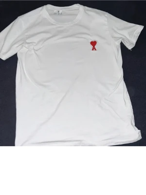Vit Ami Paris t-shirt med rött hjärta - Säljer en clean vit t-shirt från Ami Paris med det ikoniska röda hjärta-A broderiet på bröstet. Klassisk passform och rund halsringning. Tillverkad i mjuk bomull som känns skön mot huden. Perfekt för dig som gillar stilrena plagg med en twist.
