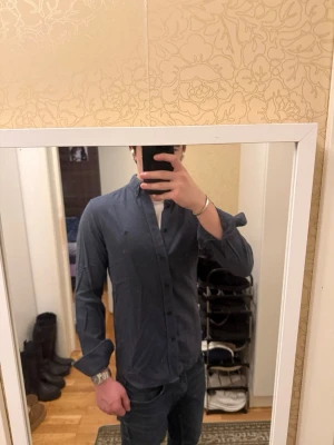 Mörkblå/grå skjorta från Morris - Säljer en stilren mörkblå skjorta från Morris i 100% bomull. Passar även S. Skjortan har klassisk krage med knappar, lång ärm och diskret logga broderad på bröstet. Perfekt för dig som gillar en clean look och vill ha något som funkar till många outfits.