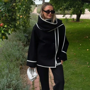 Svart kappa med vit kant - Säljer en svart kappa med oversized passform och bred, svepande krage. Jackan har vita kontrasterande kanter längs krage, ärmslut och framkant. Stängs med knappar framtill och har en modern, stilren look som sticker ut.