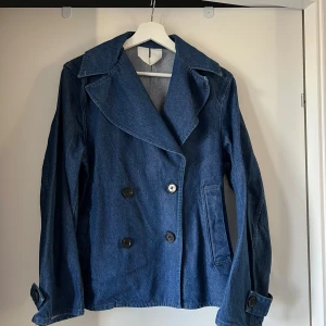 Blå jeans jacka - Säljer en snygg jacka i blått jeansmaterial med dubbelknäppning och bred krage. Jackan har långa ärmar, två framfickor och dekorativa knappar vid ärmsluten.