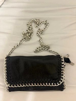 Svart axelväska med kedja från Tiamo - Säljer en svart axelväska från Tiamo i skinnimitation med grov silverfärgad kedja som axelrem och detaljer längs kanterna. Väskan har magnetlås, innerfack med dragkedja och coolt flätad kant. Perfekt för dig som gillar edgy accessoarer. Lite smutsiga för dom har varit i källare aldrig använt säljer båda för 450kr pris kan vi diskutera 