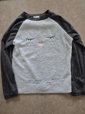 Tröja - Mysig grå fleece tröja från Cubus i storlek S. Framsidan är ljusgrå med ett broderat sött ansikte och texten 'SLEEP ALL DAY' längst ner. Ärmar och baksida är mörkgrå. Tillverkad i 100% polyester, perfekt för kalla dagar.