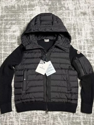 Svart cardigan från Moncler - Snygg svart cardigan från Moncler med stickade ärmar och huva. Jackan har dragkedja framtill, två fickor med knappar och en extra ficka på ärmen. Perfekt för kyliga dagar och har en modern, sportig look.