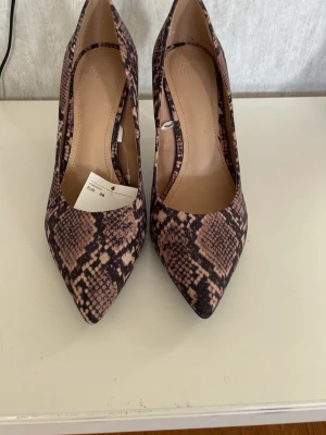 Ormskinnsmönstrade pumps från H&M - Säljer ett par snygga pumps från H&M i storlek 38. Skorna har ett ormskinnsmönster i beige och svart, spetsig tå och hög klack. Utsidan är i syntetmaterial och insidan är ljusbrun. Perfekta för dig som vill sticka ut lite extra. 