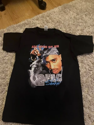 Svart 2Pac t-shirt All Eyez On Me S - Svart t-shirt med tryck av 2Pac och texten 'All Eyez On Me' på bröstet. Klassisk passform med korta ärmar och rund hals. Materialet är mjuk bomull, perfekt för dig som gillar streetwear och ikoniska motiv.