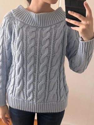 Ljusblå kabelstickad tröja - Superfin ljusblå kabelstickad tröja med bred ribbad krage och långa ärmar. Tröjan har ett chunky flätmönster och är gjord i mjukt stickat material. Perfekt för kyliga dagar och enkel att styla med jeans eller kjol.