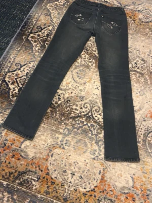 Mörkblå jeans med detaljer - Säljer ett par mörkblå jeans med raka ben och snygga broderade detaljer på bakfickorna. Jeansen har klassisk femficksdesign, silverfärgade nitar och knappar samt små dekorstenar vid fickan fram. Materialet är slitstarkt jeans. Storlek 38.