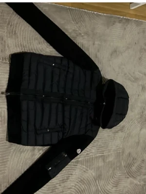 Svart Moncler pufferjacka med huva - Säljer en svart pufferjacka från Moncler med huva och dragkedja framtill. Jackan har quiltad kropp, släta ärmar och fickor med dragkedja. Moncler-logga på ena ärmen ger extra stilpoäng. Perfekt för kyliga dagar.