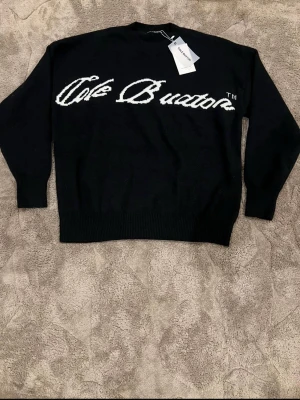 Svart stickad tröja från cole Buxto - Säljer en svart stickad tröja från  Buxton med vit logotyp i stor text över bröstet. Tröjan har rund halsringning, ribbade muddar och lång ärm. Perfekt för dig som gillar streetwear och vill ha en clean look.