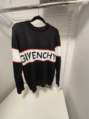 Svart stickad tröja från Givenchy - Säljer en svart stickad tröja från Givenchy med vit panel och röd kant över bröstet där loggan är invävd. Tröjan har rund halsringning och långa ärmar. Perfekt för dig som vill ha en clean men ändå statement look.