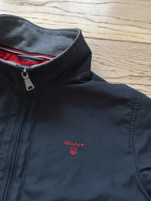 Mörkblå bomberjacka från GANT - Säljer en mörkblå bomberjacka från GANT med röd logga på bröstet och röda detaljer vid fickorna. Jackan har hög krage, dragkedja framtill och ribbade muddar. Perfekt för dig som gillar stilrena plagg.