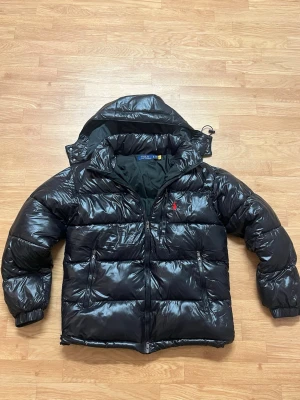 Svart pufferjacka från Polo Ralph Lauren - Säljer en svart, glansig pufferjacka från Polo Ralph Lauren i storlek M. Jackan har huva, två dragkedjefickor och den klassiska röda Polo-loggan broderad på bröstet. Perfekt för kalla dagar och riktigt snygg till streetwear-looken.
