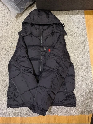 Svart pufferjacka från Polo Ralph Lauren size S - Säljer en svart pufferjacka från Polo Ralph Lauren med huva och dragkedja framtill. Jackan har det klassiska röda Polo-logot på bröstet och flera praktiska fickor med dragkedja. Perfekt för kalla dagar och riktigt snygg till streetwear-stilen.