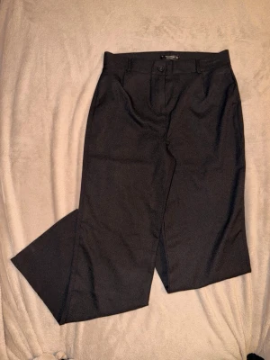 Svarta kostymbyxor från Pull&Bear - Säljer ett par svarta kostymbyxor från Pull&Bear med klassisk rak passform och hällor för bälte. Byxorna har knapp och dragkedja framtill samt diskreta fickor. Perfekta till en clean och stilren look.