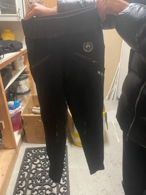 Svarta ridbyxor RH 158-164 - Säljer ett par svarta ridbyxor från RH i storlek 158-164. Byxorna har bred, elastisk midja i mörkgrått, dragkedjefickor framtill och ett litet emblem på låret. Materialet är stretchigt och passar perfekt för ridning.