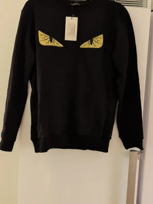 Svart sweatshirt  XL - Svart sweatshirt  i storlek XL med ikoniska gula ögon framtill. Tröjan har lång ärm och klassisk passform. Tillverkad i mjuk bomull som känns skön mot huden. Perfekt för dig som vill ha en statement piece i garderoben.